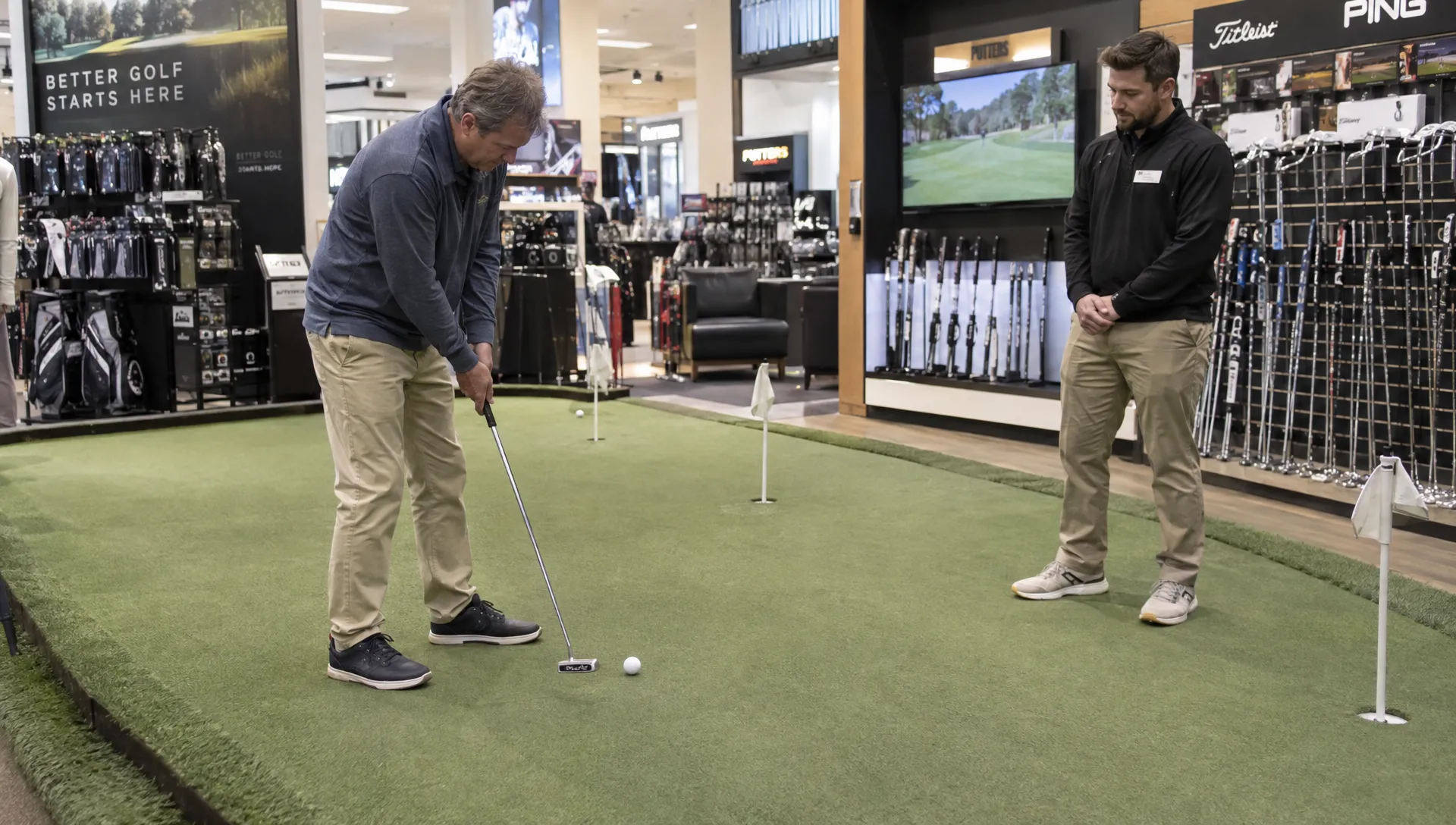 A golfer tests putts from short and medium distances on an indoor store green while a staff member watches beside the practice area.