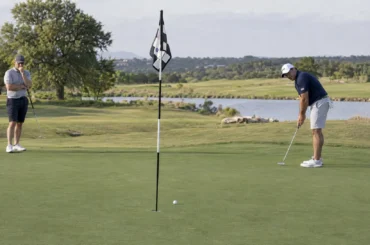 Two golfers observe a putt rolling toward the cup to judge the green's slope and pace.