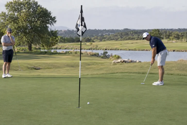 Two golfers observe a putt rolling toward the cup to judge the green's slope and pace.
